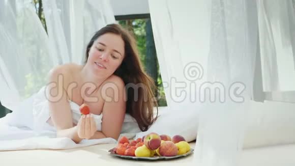 年轻的女人躺在床上手里拿着草莓在游泳池旁边的一个有窗帘的休息室里浪漫蜜月视频的预览图