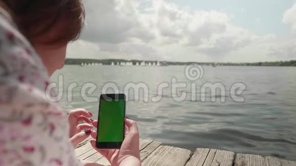 女孩躺在河边的木墩上看着绿色的智能手机屏幕用手指放大它视频的预览图