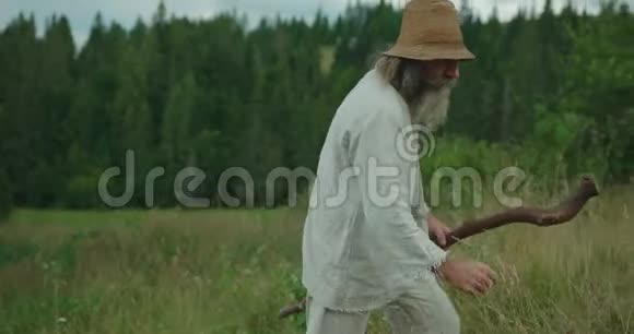 这位留着长胡子和头发的老人的特写镜头是沿着绿色的道路走的时候捡起田野的花视频的预览图