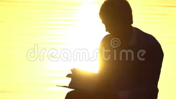 在日落时分一个金发男人在湖岸读一本书视频的预览图