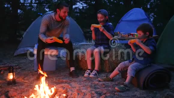 爸爸带着儿子们在暑假从外面的火堆里吃掉新鲜的黄玉米晚上在大自然的火焰附近吃晚饭视频的预览图