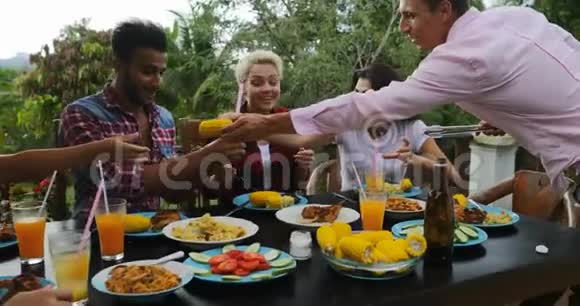 男人把食物递给坐在餐桌旁的人吃着年轻的朋友在夏天的露台上聚会视频的预览图
