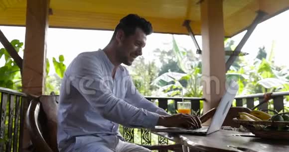 在热带森林的夏季露台酒店年轻的商人在早餐时用笔记本电脑工作小伙子一边喝果汁视频的预览图