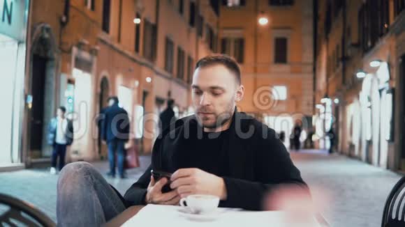 晚上年轻人坐在外面的城市咖啡馆里英俊的男性与朋友交谈使用智能手机视频的预览图