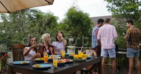 年轻女孩坐在餐桌上自拍拍照而男性团体烹饪烧烤朋友聚集在夏季露台上视频的预览图