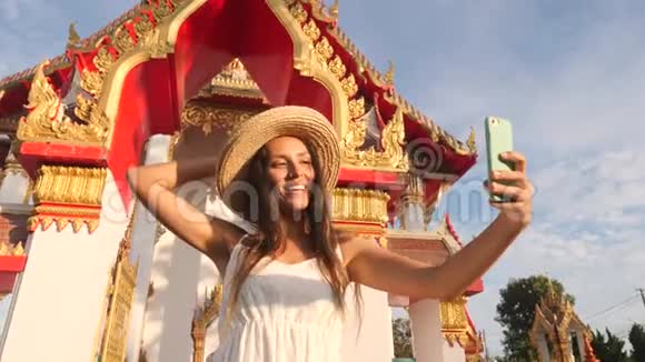 穿着白衣服戴着大草帽的年轻旅游女孩在泰国佛寺用手机拍照视频的预览图