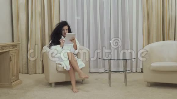 酒店客房内的扶手椅上坐着一位美丽的黑发女郎使用数字平板电脑视频的预览图