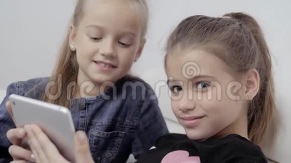 两个微笑的女孩躺在沙发上用平板电脑非常快乐的小学生的特写照片视频的预览图