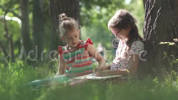 孩子们坐在树旁的草地上看一本书两个小女孩一起玩视频的预览图