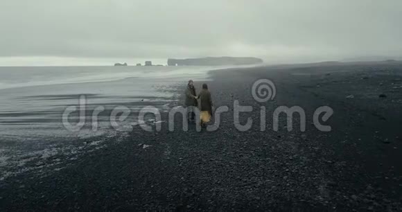 在冰岛黑色火山海滩散步的年轻时髦夫妇的背面鸟瞰图男人和女人享受大海视频的预览图
