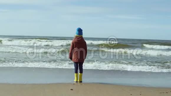 孤独的小女孩穿着明亮的黄色橡胶靴站在沙滩上休息可爱的雌性看着海浪视频的预览图