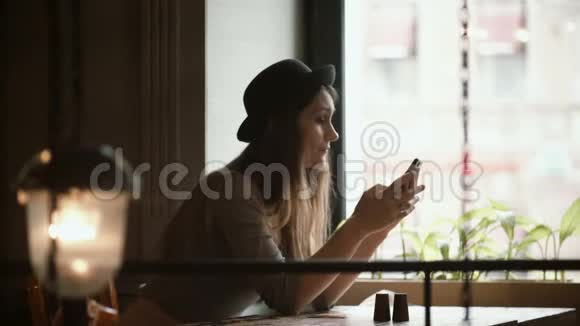 时尚的年轻女性戴着帽子在靠近窗户的咖啡馆里使用智能手机有魅力的女性上网视频的预览图