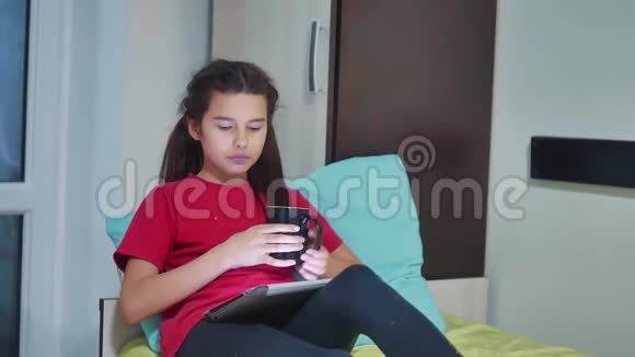 小女孩在玩数字平板电脑吃三明治小女孩坐在床上玩数字平板电脑有零食视频的预览图