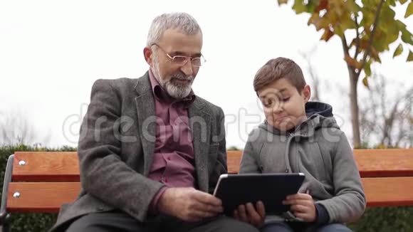 爷爷和孙子一起在公园里度过时光他们坐在长凳上用平板电脑视频的预览图