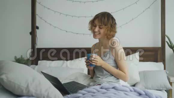 一位性格开朗的年轻女性在家里坐在床上与朋友用笔记本电脑聊天视频的预览图