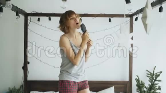 迷人的快乐的女人在床上跳舞在家里用梳子像麦克风唱歌视频的预览图