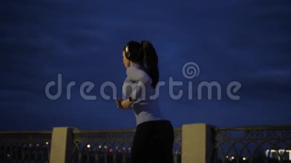 女孩用耳机听音乐晚上在城里慢跑穿着运动服的女运动员的背景视频的预览图