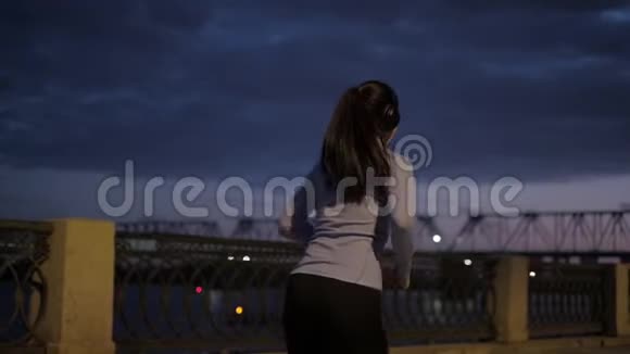 女孩用耳机听音乐晚上在城里慢跑穿着运动服的女运动员的背景视频的预览图