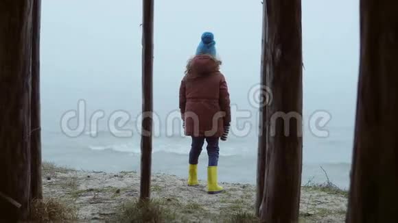 小女孩穿着橡胶靴站在海边望着海浪雾天和寒冷的日子里的孩子视频的预览图