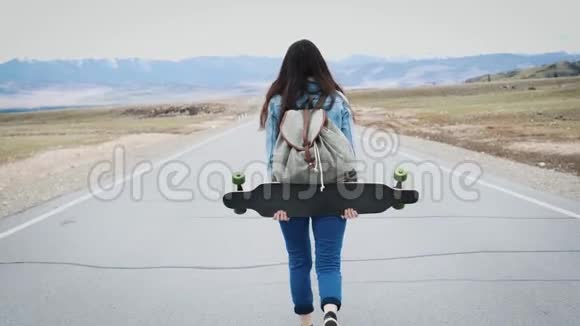 年轻女孩十几岁的嬉皮士用长板在高速公路上行走带着露营背包和运动衫反对视频的预览图