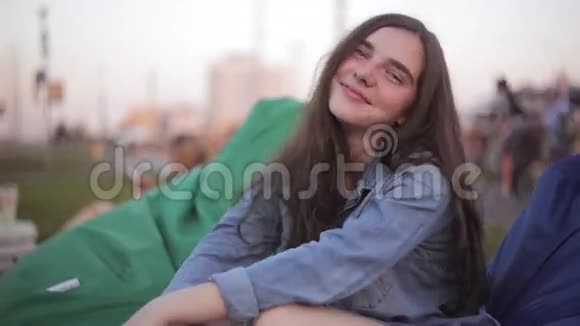 在炎热的夏天穿着牛仔裤夹克的年轻漂亮的少女在公园里微笑坐在蓝豆包椅上视频的预览图