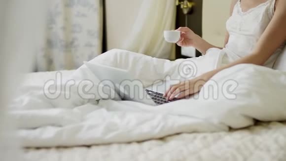 年轻的美女在床上一边喝咖啡一边用笔记本电脑视频的预览图