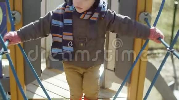 可爱的小男孩穿着橙色的橡胶靴在操场上散步孩子小心翼翼地在儿童索道上迈出第一步视频的预览图