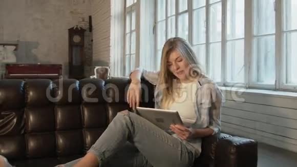 穿着破牛仔裤的漂亮金发女郎微笑着坐在沙发上通过平板电脑与父母交流一个年轻的学生视频的预览图