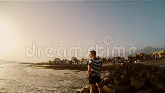 在刮风的日子里年轻的游客站在海边男性看着美丽的日落视频的预览图