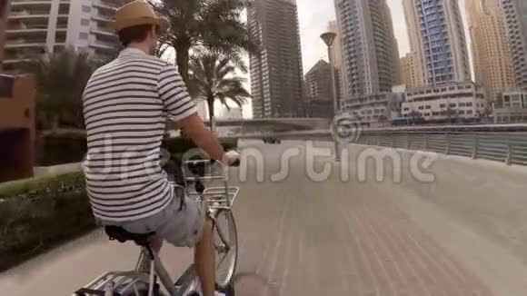 一个穿着帽子条纹T恤和短裤的年轻人骑着自行车在摩天大楼和游艇之间的长廊上视频的预览图