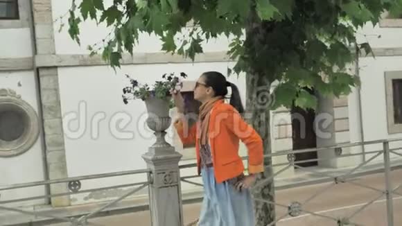 穿着橙色夹克和蓝色牛仔裤裙子的漂亮女孩在白色的街锅里闻着漂亮的花视频的预览图