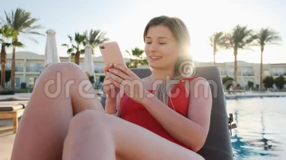 年轻性感的女人在游泳池边晒太阳用手机穿着红色比基尼的女孩躺在躺椅上视频的预览图