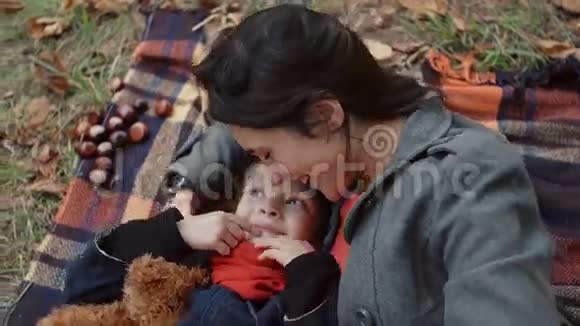 妈妈和她可爱的小女儿躺在格子布上妈妈在秋天公园的鼻子里挠痒痒和亲吻女孩视频的预览图