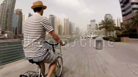 一个穿着帽子条纹T恤和短裤的年轻人骑着自行车在摩天大楼和游艇之间的长廊上视频的预览图