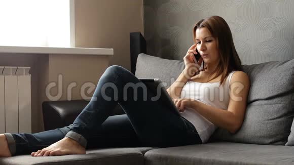 女人一边笑一边在电话里说话用平板电脑女人用现代电子设备坐在沙发上视频的预览图