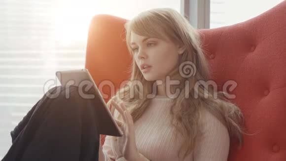 年轻女子在家坐在窗前的红色扶手椅上用平板电脑工作视频的预览图