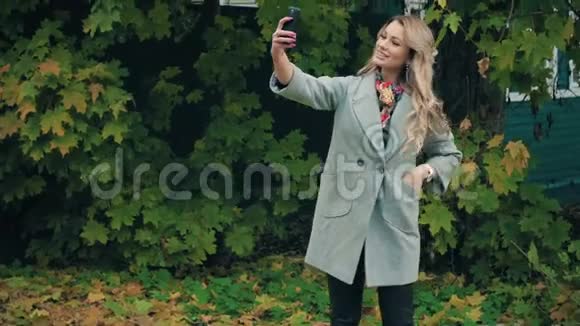 穿着灰色外套的金发美女在秋日公园的智能手机上自拍视频的预览图