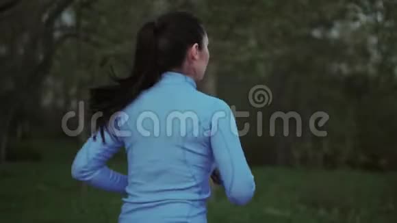 慢跑一个穿蓝色运动服的年轻女孩正沿着傍晚的公园跑步背面视图视频的预览图