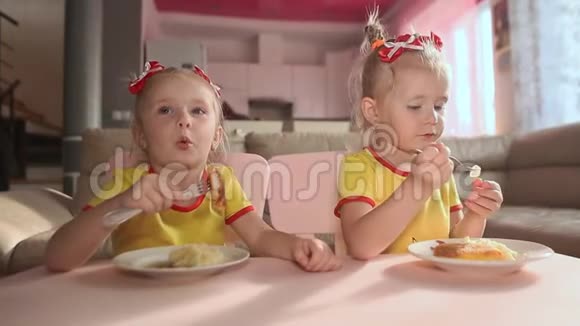两个穿着黄色T恤的小可爱双胞胎姐妹正一起在房子厨房的桌子上吃食物视频的预览图