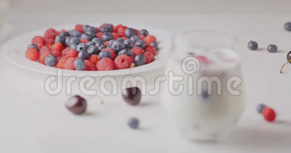 将焦点从一个带有成熟甜树莓蓝莓的盘子中转移樱桃放在一个带有浆果和牛奶的杯子上放在白色的桌子上视频的预览图