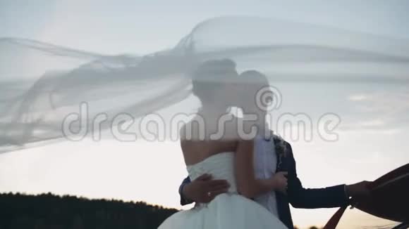 面纱迎风飘扬新娘和新郎在日落时温柔地拥抱婚礼后美丽的男人和女人的夜晚视频的预览图