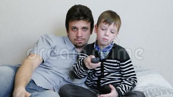 在沙发上看电视无聊的男孩和他爸爸正在看电视并切换频道视频的预览图