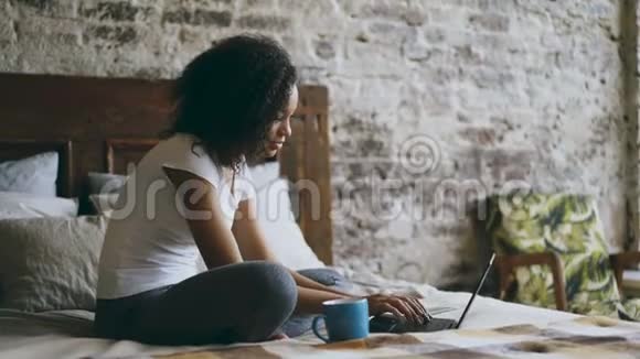 可爱的非洲女孩在笔记本电脑上打字以便在家里的床上分享社交媒体视频的预览图