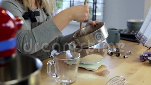 年轻女子站在厨房里用面粉在碗里筛可可金发女性称重配料视频的预览图