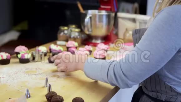 年轻的金发女人用彩色奶油装饰纸杯蛋糕用糕点袋烹调的漂亮糖果视频的预览图