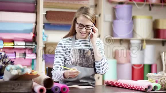 一个年轻的女人正在花店里工作她的笔记本电脑和电话快乐的花艺女人用纸包花视频的预览图