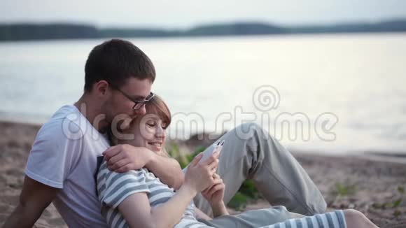 年轻漂亮的夫妇坐在海滩上在河岸上聊天女人用智能手机视频的预览图