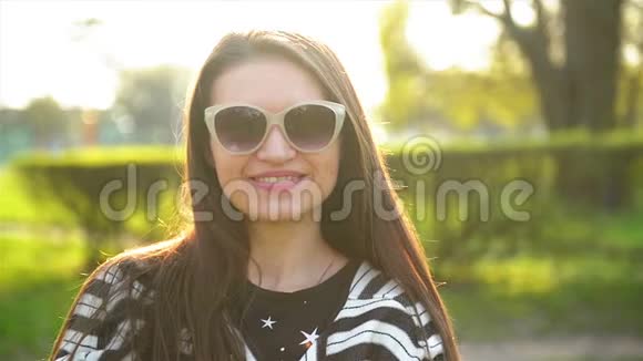 在阳光明媚的春天里为一位身穿太阳镜和条纹衬衫的漂亮黑发女郎特写户外肖像视频的预览图