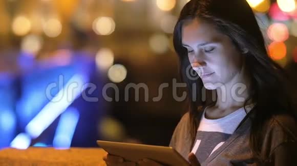 一个美丽的黑发女人微笑着用她的平板电脑夜晚的城市背景上模糊的灯光慢悠悠视频的预览图