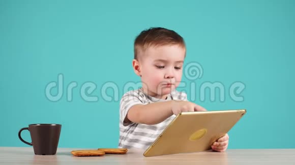 可爱的小男孩用平板电脑坐在桌子上孤立在蓝色上视频的预览图